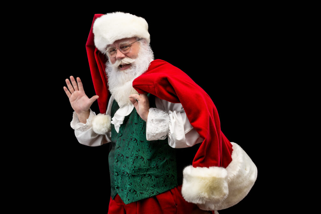 Santa Jim of Virginia Santa Claus smiles and waves with his right hand while wearing a green vest, white ruffled shirt and red hat with white fur trim. H is red jacket with white fur trim is draped over his left shoulder.